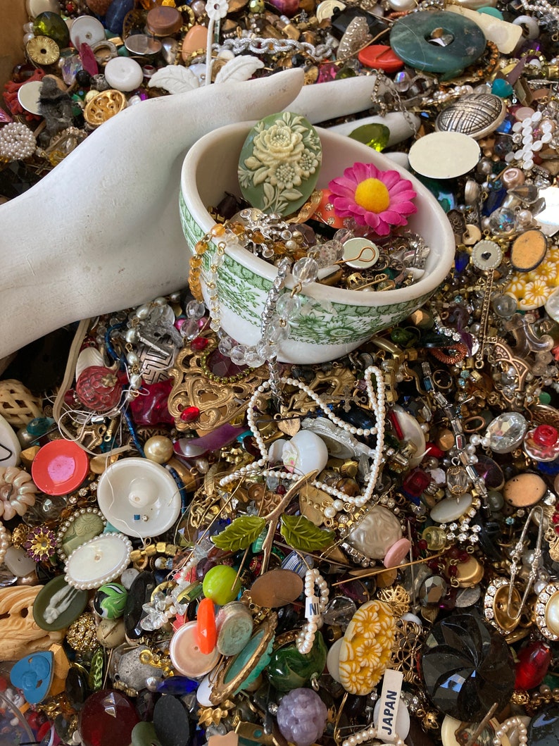 May include: A white ceramic bowl filled with a variety of colorful buttons, beads, and jewelry. The bowl is sitting on a table covered in a large collection of buttons, beads, and jewelry.