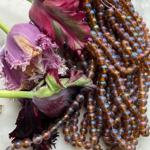 May include: Close-up of a collection of brown iridescent glass beads, strung together, with a cluster of dark purple and pink tulips. The tulips have ruffled petals with green accents. The beads have a shimmering, oil-slick effect.