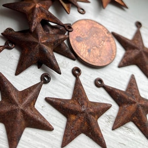 May include: A collection of rustic, star-shaped metal ornaments with a distressed, rusty brown finish. Each star has a small loop for hanging. A copper penny is also visible, providing a size reference. These decorative stars are ideal for crafting or holiday decor.