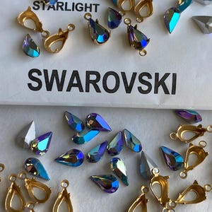 May include: A close-up of a variety of iridescent blue teardrop-shaped crystals and gold-toned metal settings. The crystals are labeled "Swarovski" and "Starlight".