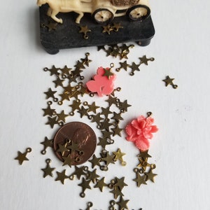 K&ouml;nnte beinhalten: Eine gro&szlig;e Auswahl an goldenen sternf&ouml;rmigen Charms mit Sprungringen. Die Charms sind auf einer wei&szlig;en Oberfl&auml;che mit einem Penny und zwei rosa Plastikblumen verstreut.