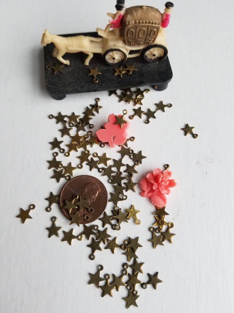 K&ouml;nnte beinhalten: Eine gro&szlig;e Auswahl an goldenen sternf&ouml;rmigen Charms mit Sprungringen. Die Charms sind auf einer wei&szlig;en Oberfl&auml;che mit einem Penny und zwei rosa Plastikblumen verstreut.