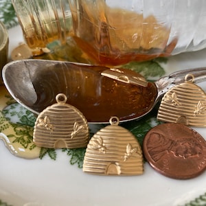 May include: Close-up of gold-colored beehive charms with bee details, a penny, and a spoon with a honey-like substance. The charms are on a white plate with a green floral pattern. A glass jar is in the background.