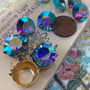 May include: An assortment of iridescent, faceted rhinestones in various shapes and sizes, including round and oval. A gold-colored metal jewelry finding is also present. The rhinestones display a range of colors, including blue, green, and purple.