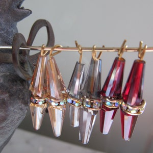 May include: Six pairs of dangle earrings with crystal teardrop shapes. The earrings are gold-toned and have a clear, iridescent finish. The teardrops are adorned with a row of sparkling rhinestones.
