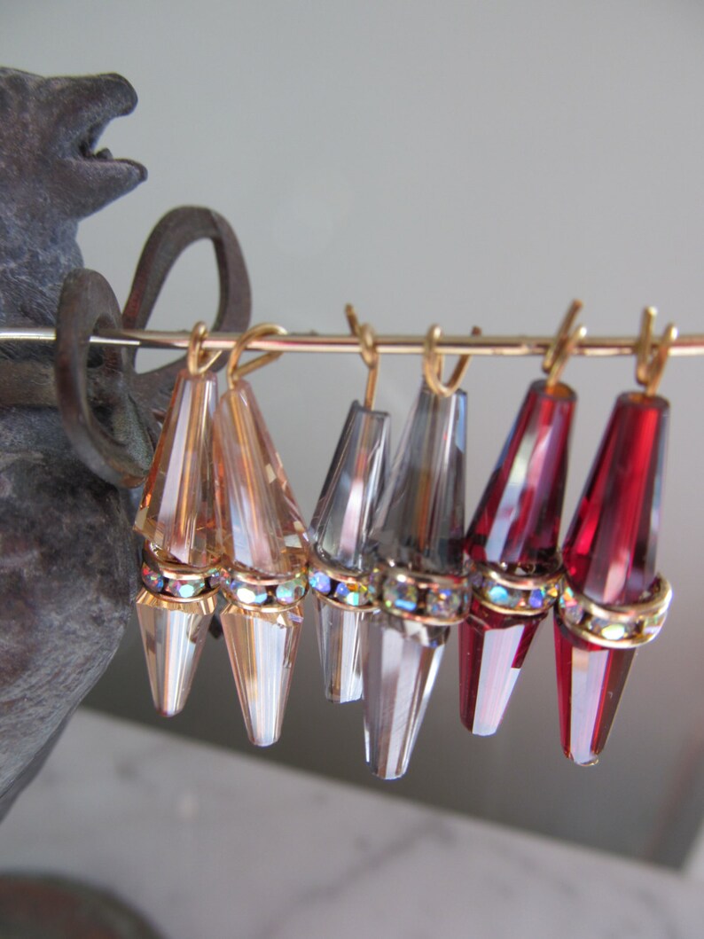 May include: Six pairs of dangle earrings with crystal teardrop shapes. The earrings are gold-toned and have a clear, iridescent finish. The teardrops are adorned with a row of sparkling rhinestones.