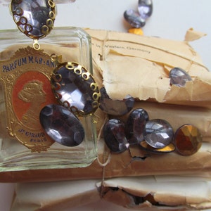 May include: A vintage glass bottle of perfume with a gold cap and a label that reads "Parfum Marian J. Giraud Paris". The bottle is surrounded by a variety of small, clear glass stones with gold accents.