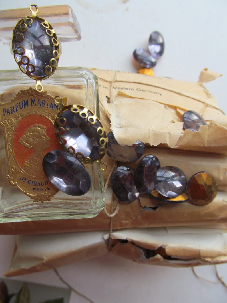 May include: A vintage glass bottle of perfume with a gold cap and a label that reads "Parfum Marian J. Giraud Paris". The bottle is surrounded by a variety of small, clear glass stones with gold accents.