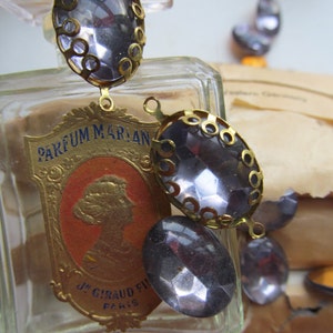 May include: A clear glass bottle with a gold label that reads "PARFUM MARIAN J. GIRAUD FILS PARIS". The bottle is decorated with a gold chain and several clear glass stones.