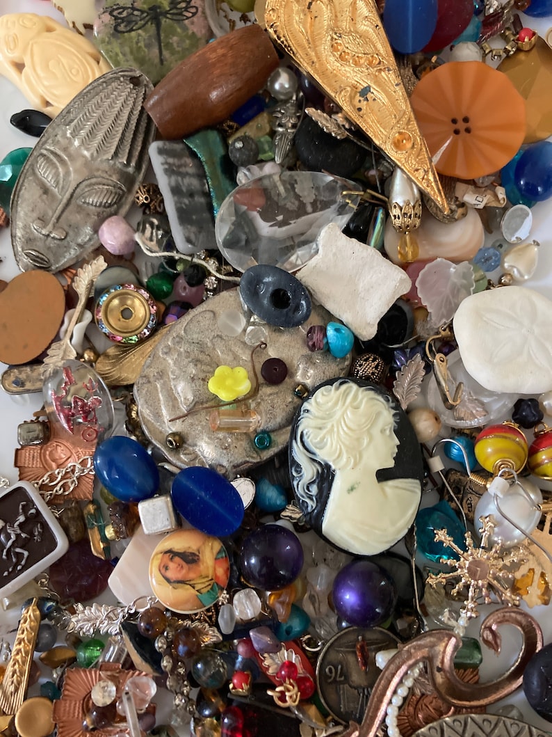 May include: A collection of colorful and eclectic jewelry making supplies, including beads, buttons, charms, and findings. The supplies are arranged in a random pattern on a white surface.