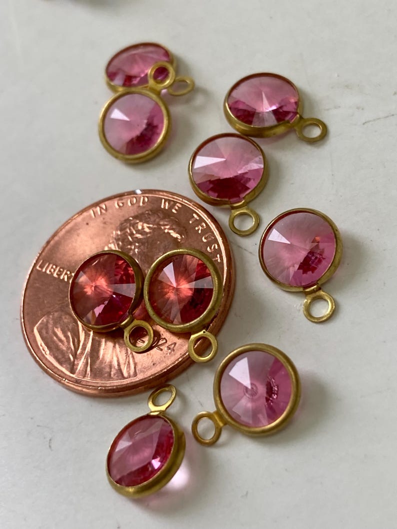 May include: A collection of pink gemstone pendants with gold-colored metal settings. The faceted stones vary in size and shape, with a small loop at the top for attaching to jewelry. A penny is included for size reference.