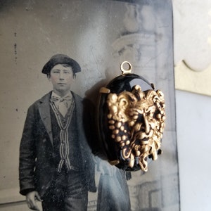 May include: A gold-toned pendant with a black cameo featuring a detailed, grotesque face with a beard and grapes. The cameo is set in a gold-toned frame with a loop for hanging.