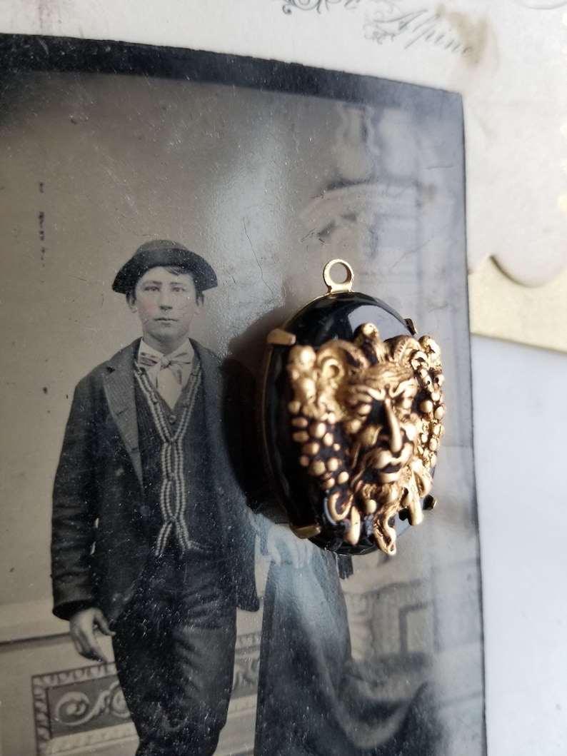 May include: A gold-toned pendant with a black cameo featuring a detailed, grotesque face with a beard and grapes. The cameo is set in a gold-toned frame with a loop for hanging.