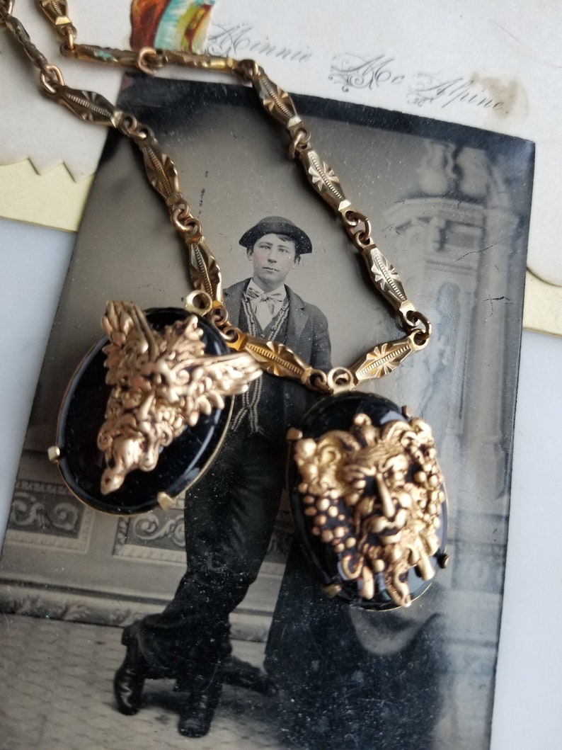 May include: A gold chain necklace with two black cameo pendants featuring gold-toned, detailed faces. The pendants are oval-shaped and have a textured, almost 3D effect. The chain is ornate and has a delicate, intricate design.