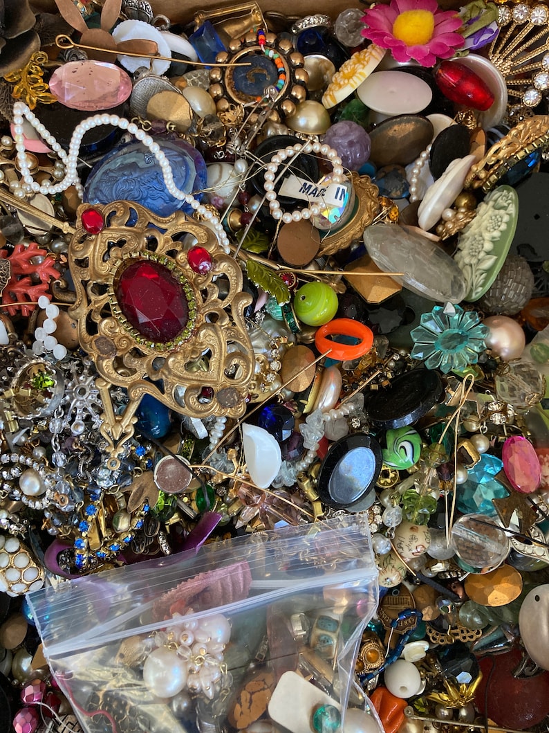 May include: A large collection of colorful and assorted buttons, beads, and jewelry findings. The items are arranged in a box and include a variety of materials, shapes, and sizes. There is a small plastic bag in the lower right corner of the image.