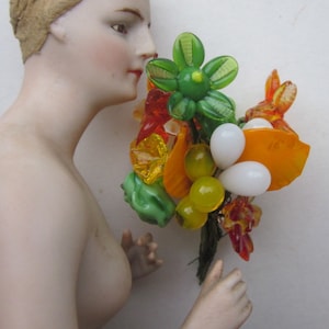 Peut inclure: Un bouquet de perles de verre colorées dans des tons de vert, orange, jaune et blanc. Les perles sont disposées en forme de fleur avec un centre vert et des perles blanches qui l'entourent. Le bouquet est tenu par une poupée en porcelaine.