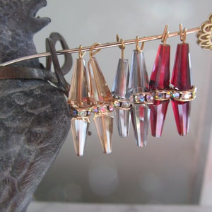 May include: A set of six teardrop-shaped crystal earrings with gold-tone tops. The earrings are arranged in a row on a metal stand. The earrings are clear, silver, and red.