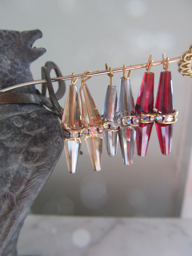 May include: A set of six teardrop-shaped crystal earrings with gold-tone tops. The earrings are arranged in a row on a metal stand. The earrings are clear, silver, and red.
