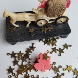 K&ouml;nnte beinhalten: Eine Miniatur-Kutsche aus wei&szlig;em Material, die von einem wei&szlig;en Pferd gezogen wird. In der Kutsche befinden sich zwei Figuren. Die Kutsche ist mit goldenen Verzierungen verziert und die Figuren tragen rosa und schwarze Kleidung. Die Kutsche steht auf einem schwarzen Sockel, um den herum goldene Sterne verstreut sind.