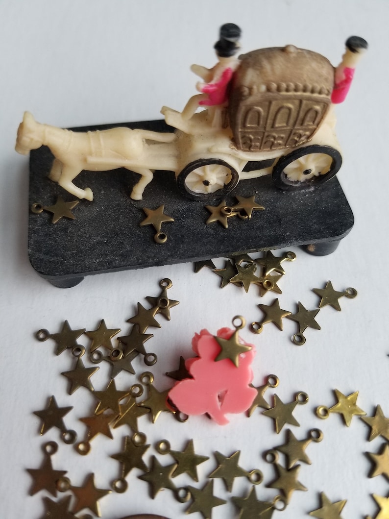K&ouml;nnte beinhalten: Eine Miniatur-Kutsche aus wei&szlig;em Material, die von einem wei&szlig;en Pferd gezogen wird. In der Kutsche befinden sich zwei Figuren. Die Kutsche ist mit goldenen Verzierungen verziert und die Figuren tragen rosa und schwarze Kleidung. Die Kutsche steht auf einem schwarzen Sockel, um den herum goldene Sterne verstreut sind.