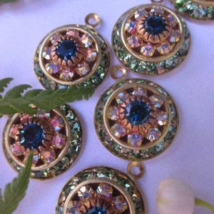 May include: Five gold-toned pendants with a round, multi-faceted, iridescent crystal center surrounded by smaller crystals. The crystals are in shades of blue, pink, and green. The text "Peridot Crystal AB Indicolite" is visible at the top of the image.