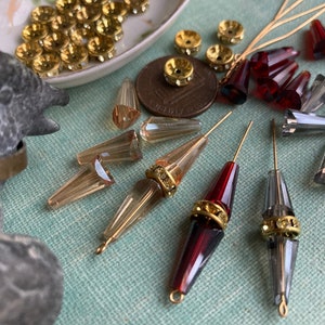 May include: Assortment of vintage glass beads and gold-toned metal beads with rhinestones. The beads are in various shapes and sizes, including teardrop, round, and conical. The beads are arranged on a blue surface.