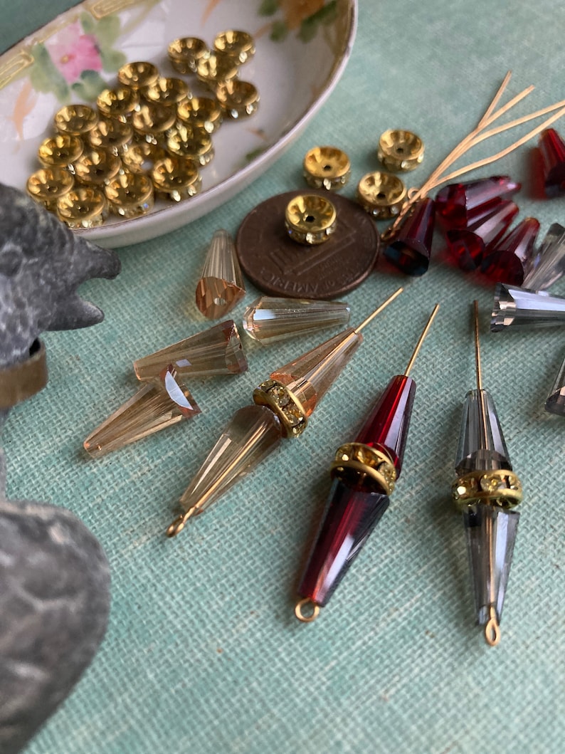 May include: Assortment of vintage glass beads and gold-toned metal beads with rhinestones. The beads are in various shapes and sizes, including teardrop, round, and conical. The beads are arranged on a blue surface.