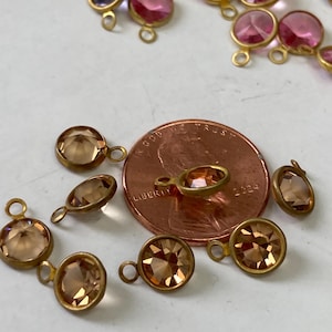 May include: Close-up of various gemstone charms in shades of pink, purple, and amber, each with a gold-colored metal setting and a small loop. A penny is included for size reference.