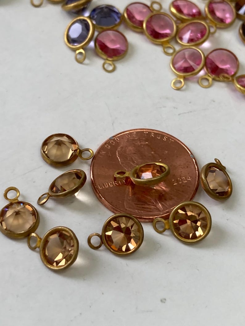 May include: Close-up of various gemstone charms in shades of pink, purple, and amber, each with a gold-colored metal setting and a small loop. A penny is included for size reference.