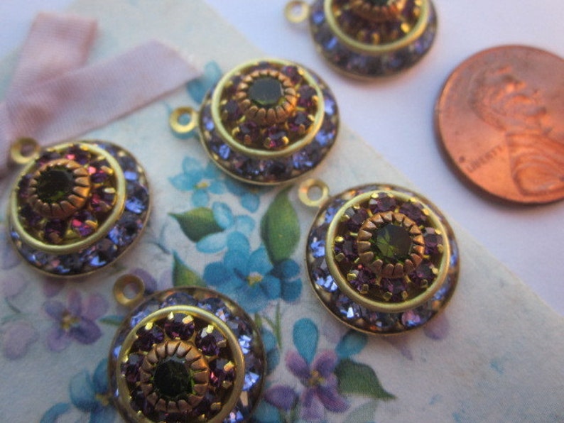 May include: Five gold-toned metal charms with a round, multi-layered design. Each charm features a central green gemstone surrounded by smaller purple gemstones. The charms have a loop at the top for attaching to jewelry or other crafts.