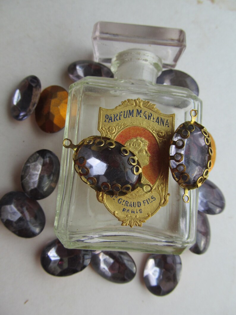May include: A vintage glass bottle with a gold label that reads "PARFUM MARLANA GIRAUD FILS PARIS". The bottle is surrounded by small, purple glass stones with gold accents.