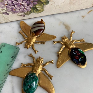 May include: Three gold bee-shaped ornaments with glass accents. The bees have detailed wings and bodies. One bee has a brown and white striped glass accent, one has a green glass accent, and one has a black and red glass accent.