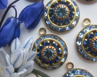 May include: Close-up of several round, ornate pendants with a central blue gemstone surrounded by smaller blue crystals. The pendants have a gold-colored metal frame and a loop at the top. Blue flowers are in the background.