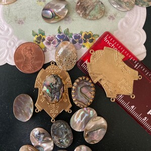May include: An assortment of iridescent abalone shell cabochons and gold-toned metal findings. A red ruler with inch measurements and a U.S. penny are also present. The shells vary in size and shape, with a range of colors including pearlescent, brown, and gray.