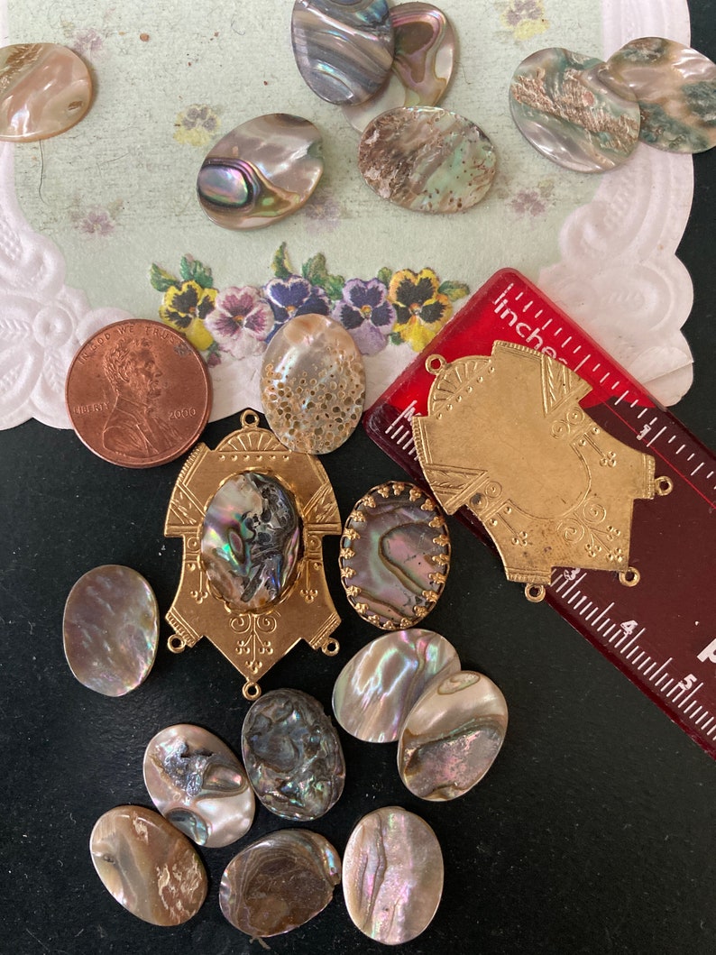May include: An assortment of iridescent abalone shell cabochons and gold-toned metal findings. A red ruler with inch measurements and a U.S. penny are also present. The shells vary in size and shape, with a range of colors including pearlescent, brown, and gray.