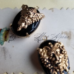 May include: Two black cameo pendants with gold-toned metalwork depicting a face with a beard and a crown. The pendants are oval shaped and have a black background.