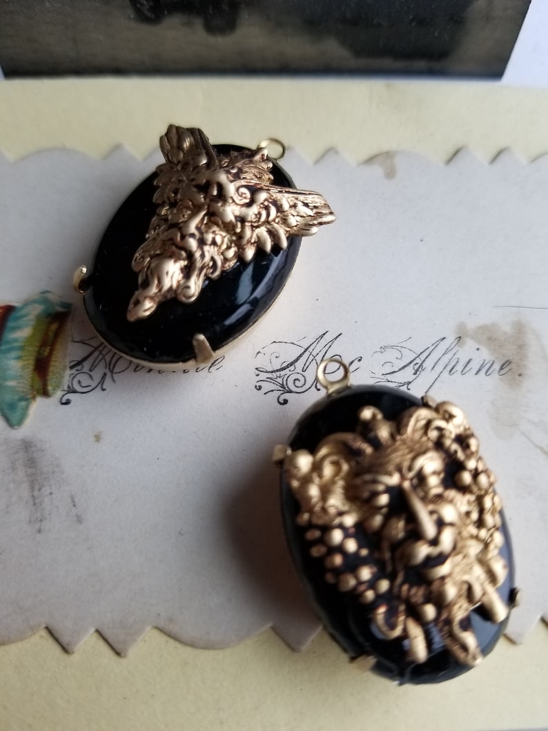 May include: Two black cameo pendants with gold-toned metalwork depicting a face with a beard and a crown. The pendants are oval shaped and have a black background.