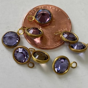 May include: A collection of small, round and oval purple gemstone charms with gold-colored metal settings. Each charm has a loop for attaching to jewelry. The gemstones vary in shade from light to dark purple.