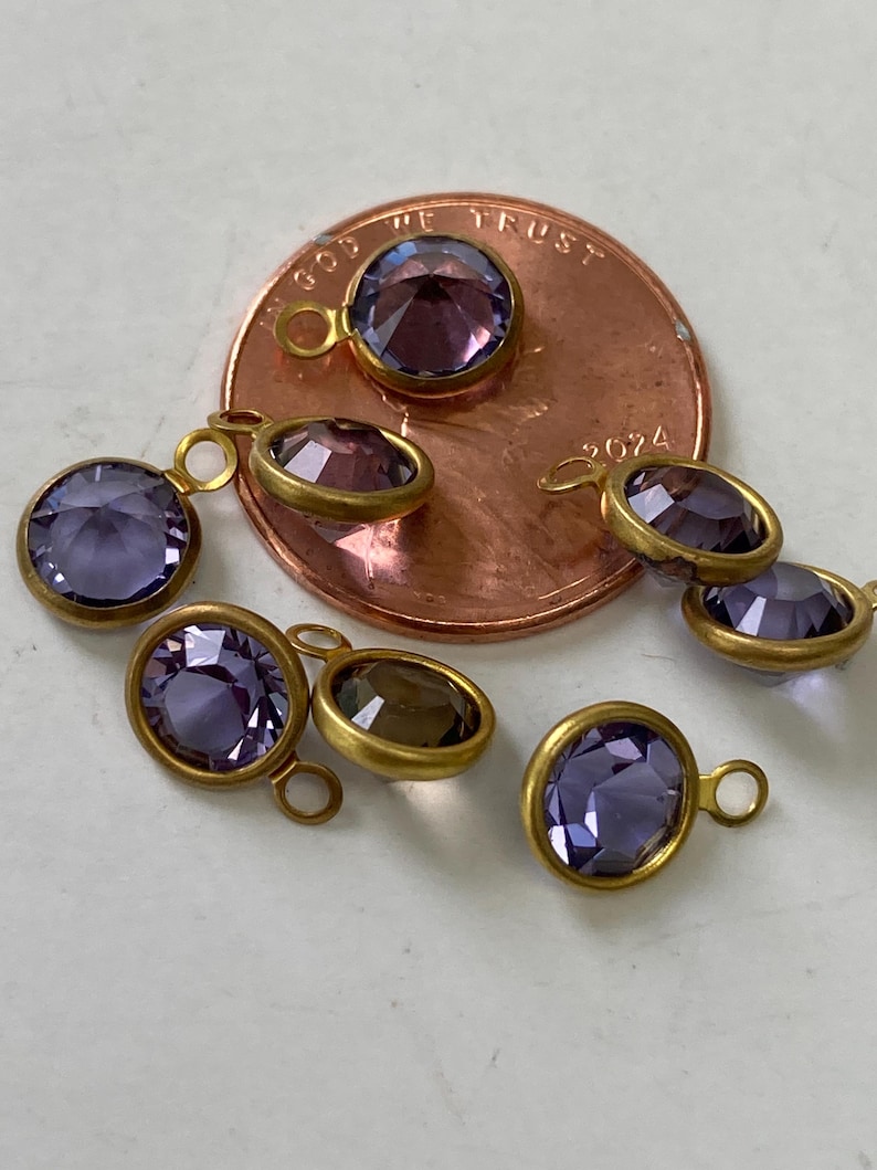 May include: A collection of small, round and oval purple gemstone charms with gold-colored metal settings. Each charm has a loop for attaching to jewelry. The gemstones vary in shade from light to dark purple.