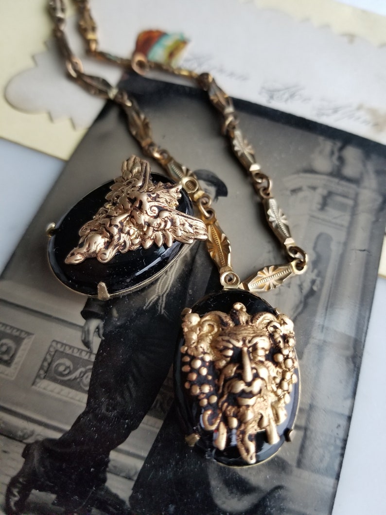 May include: A gold chain necklace with two black cameo pendants featuring detailed gold sculpted faces with foliage accents.