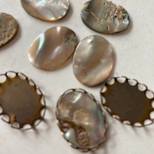 May include: A collection of iridescent oval-shaped mother-of-pearl cabochons and decorative metal settings. The cabochons display a range of colors, including white, tan, and hints of blue and green. Some are set in ornate metal frames.