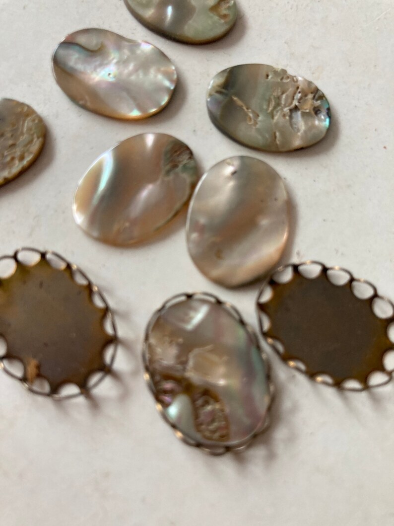 May include: A collection of iridescent oval-shaped mother-of-pearl cabochons and decorative metal settings. The cabochons display a range of colors, including white, tan, and hints of blue and green. Some are set in ornate metal frames.