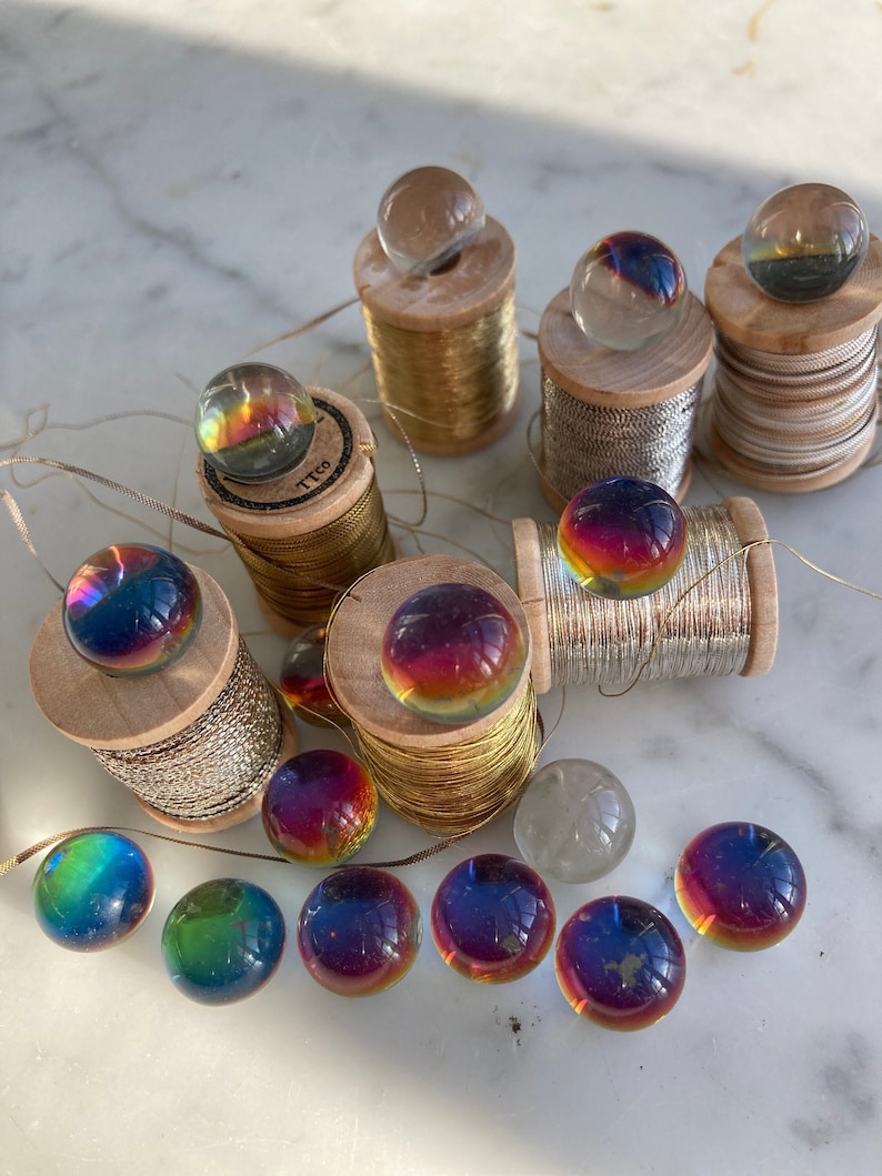 Peut inclure: Une collection de billes de verre iris&eacute;es dispos&eacute;es sur des bobines de fil. Les billes sont dispos&eacute;es en cercle autour des bobines, certaines &eacute;tant plac&eacute;es sur les bobines. Les bobines sont en bois et de diff&eacute;rentes couleurs, notamment or, argent et blanc.