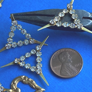 May include: Close-up of gold-toned jewelry components, including rhinestone-studded triangles and a heart-shaped piece. A penny from 1978 is included for scale. The background is a solid blue.
