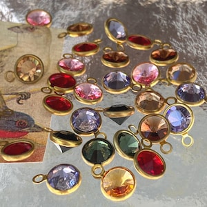 May include: A collection of colorful gemstone pendants with gold-colored settings. The gemstones are in various shapes and colors, including pink, red, purple, and amber. Each pendant has a small loop for attaching to a chain or other jewelry component.