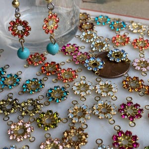 May include: Close-up of floral earrings and decorative jewelry components. The earrings feature red and orange crystals with turquoise beads. Various flower-shaped components with colorful crystals are scattered on a white surface.