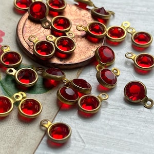 May include: A collection of small, red, round gemstone charms with gold-colored metal settings and loops. The charms are scattered on a light-colored surface, with a penny visible in the background.