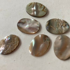 May include: Six oval-shaped, iridescent mother-of-pearl cabochons. The cabochons have a pearlescent sheen with variations of beige, gray, and hints of blue and green. The surfaces have natural textures and imperfections.