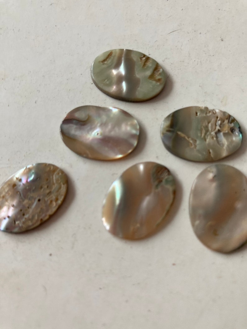 May include: Six oval-shaped, iridescent mother-of-pearl cabochons. The cabochons have a pearlescent sheen with variations of beige, gray, and hints of blue and green. The surfaces have natural textures and imperfections.