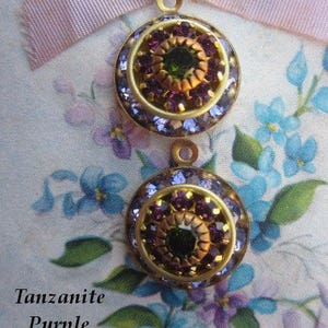 May include: Two gold-toned round pendants with a center cluster of purple and green rhinestones. The pendants have a textured surface and are surrounded by a ring of smaller purple rhinestones.